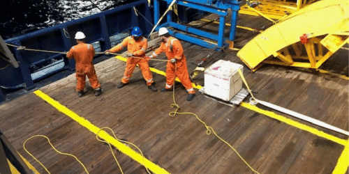 Three workers in orange coveralls and helmets handling ropes on a ship deck